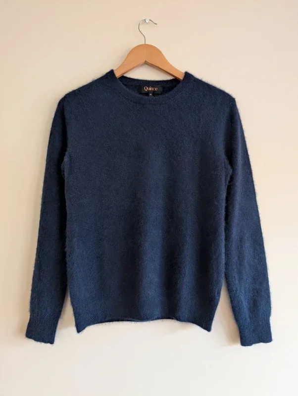 A navy Quince Mongolian cashmere crewneck sweater on a wooden hanger against a pale wall.